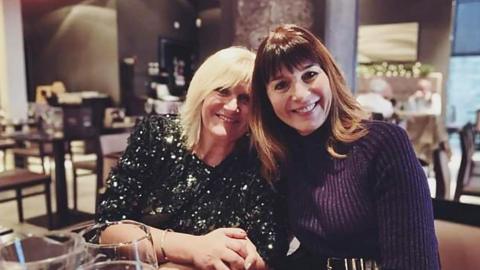 Two women, one wearing a sparkly green top and the other wearing a purple top, sit closely together in a restaurant