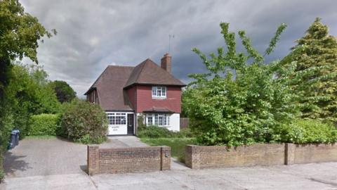 The outside of Rainscombe House care home which is a detached house with a tiled first floor and sloping roof. It has a driveway, a lawn and is surrounded by shrubs and trees.