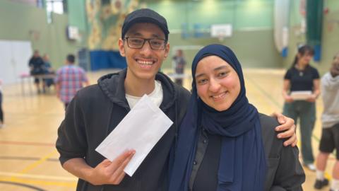 Kareem has his arm around Jana. He is holding his GCSE results envelope. They are both smiling and are in a school sports hall