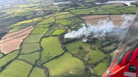 Aerial photo of a fire in a field as smoke billow across numerous green and brown fields, with a river in the background. A red corner of an aircraft obscures the bottom right corner of the frame.