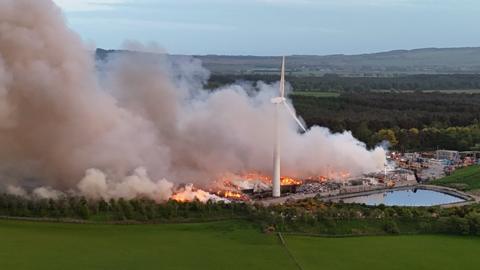 A landfill site next to some water and a windmill is on fire with smoke rising from the scene.