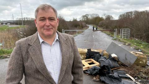 Philip Spicer with short brown hair, wearing a brown jacket over a white shirt. He is standing beside some rubbish dumped in front of a gated road. It includes black-bagged waste, pieces of wood and tree branches