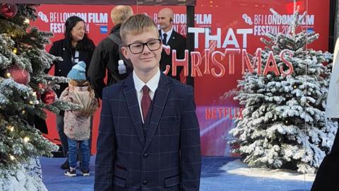 Jack on the red carpet release of That Christmas. He has light coloured hair and is wearing a suit and black glasses. 