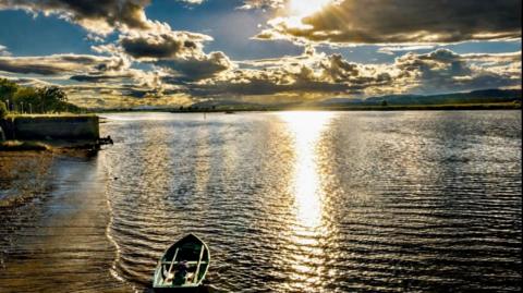 Rays of sunshine hit the water despite mostly dark clouds in the sky. A small rowing boat is sitting in the bay. 
