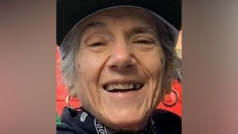 Pepita Sakirecili, also known as Pepita Garcia, wearing a cap and gold hoop earrings laughing toward the camera. She is wearing a patterned scarf and dark top, and has grey curly hair, either worn short or tied back.