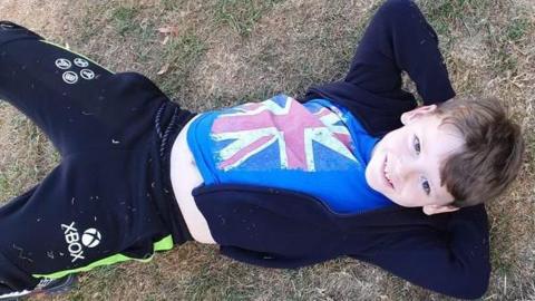 Lennix Sutcliffe. He is lying on the grass, with the picture taken from above. His hands are up behind his head and he is smiling at the camera.