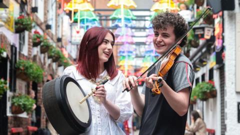 A woman and man standing in Belfast's Cathedral Quarter. The woman, with dark red hair, is holding a bodhrán. The man, with short, curly brown hair, is holding a fiddle.