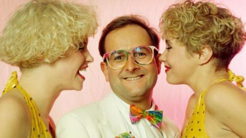 Timmy Mallett in his trademark multi-coloured glasses with a colourful bow tie stands between to blonde-haired women wearing polka-dotted bikinis