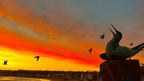 A bird statue is in silhouette against a red and yellow sky. There is water below.