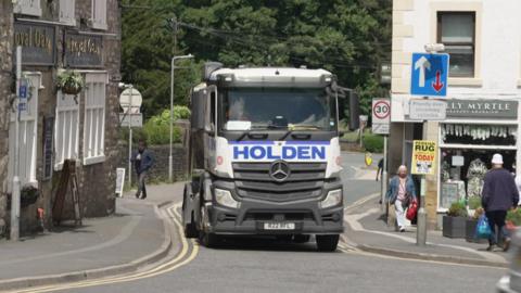 A heavy goods vehicle passes between two buildings on a narrow section of road in the town of Settle 