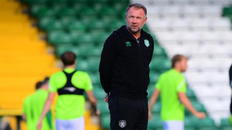 Marcus Stewart is standing on a pitch in a black outfit. Behind him, players are training in high-vis, green shirts. The bleachers are blurry in the background.