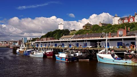 General view of North Shields' Fish Quay. A number of small boats are berthed along the dock. Clifford's Fort - a white tower - can be seen in the background.