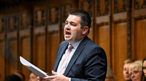 MP Shaun Davies standing in the House of Commons, wearing a blue suit and pink tie with notes in his hands