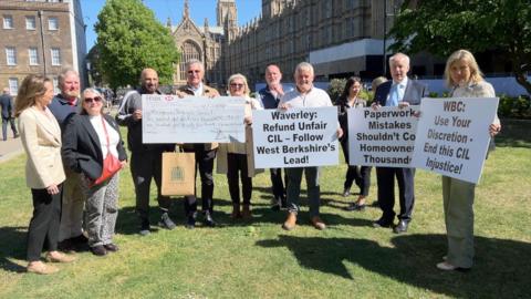 A group of people stand in front of the Houses of Parliament holding protest placards about the Cil levy in bright sunshine.