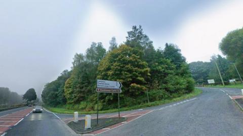 The Blenkinsopp Castle junction on the A69, close to where the fatal crash occurred. The A69 ploughs a straight course into the distance and has light traffic on it. Cars have their lights on as it is an overcast day and there is evidence of light rain. A road leads off to the right. It travels uphill then has a steep curve to the left which takes it behind a thick crop of mature trees. A white and black road sign points the way (with distances) to - from top to bottom - Lambley, Featherstone and Blenkinsopp Castle. Below that is a brown sign pointing to Blenkinsopp Castle Inn.