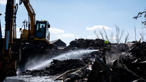 A firefighter hosing down piles of industrial waste while a mechanical digger stands by