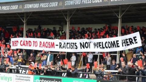A large flag which reads "Red card to racism", racists not welcome here" being held up in a football stand. Other fans are holding large red cards up. 
