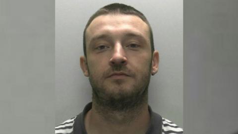 A police mugshot of William James looking directly at the camera. He is wearing a grey and white striped T-shirt with a grey collar and he has close-cropped brown hair and stubble. He has a gold earring in one ear.