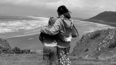 A black and white picture of Corrine Cope with her arm round Dylan. They have their backs to the camera, with Dylan wearing his hood up, and are looking out over a coastal scene