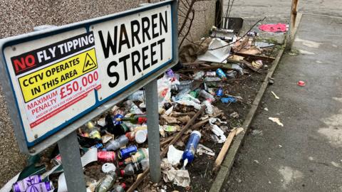 The Warren Street road sign has a sticker on it saying 'No Fly Tipping'. Beneath it are scores of empty cans and bottles.