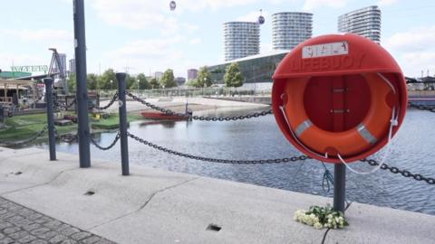 Flowers placed beneath a lifebuoy on the edge of Royal Victoria Dock. Beside the water is a boat parked by a man-made beach which is next to City Hall.
