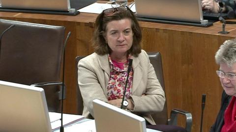 Eluned Morgan is pictured with her arms crossed wearing a white jacket sat in a chair in the debating chamber of the Welsh Parliament. Her colleague Jane Hutt  is sat next to her. They are listening to Andrew RT Davies, Welsh Conservative Senedd leader, who is not in the shot.