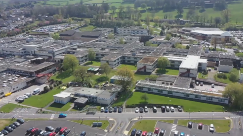 An aerial shot of grey concrete hospital building blocks and some parked cars and green patches of grass