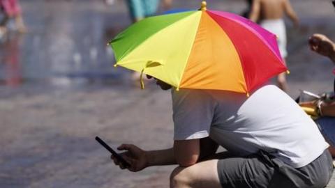 A man in a white tshirt and shorts sheltering under a coloured umbrella and looking at a mobile phone