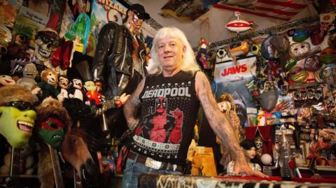 Martin leans against a red pool table surrounded by a dense collection of pop-culture memorabilia. The room contains masks, stuffed toys, posters, model displays and a life-sized figure dressed in black standing behind them.