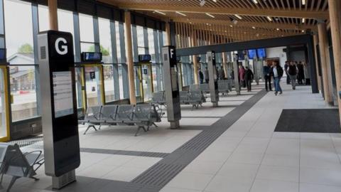 A row of bus stands inside the new Bishop Auckland bus station. There is seating and a digital screen at each stand. A group of people is walking down the hall.