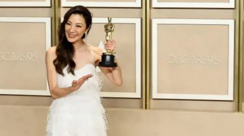 Michelle Yeoh holds up her Oscar in 2024. Her long, black hair is swept to one side and she's wearing a white dress. 