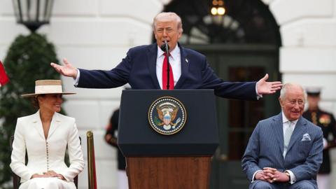 Donald Trump is standing at a lectern featuring the official coat of arms of the US presidency. He has both arms raised out to the sides. King Charles is sitting at one side and Trump's wife Melania is on the other. The King is wearing a light blue pin-striped suit with a light shirt and a light blue tie with a light blue pocket square. He is looking to the side and laughing. Trump is wearing a dark blue suit jacket, white shirt and red tie. He is mid speech. Melania is wearing a white fitted suit and a straw hat. They are outside the White House