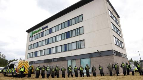 About 20 police officers in riot gear lined up in front of the multi-storey hotel. A police van can be seen to the left.