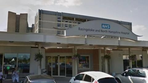 A view of the front entrance of Basingstoke and North Hampshire Hospital. The building is a faded cream colour, and there are cars parked outside the front entrance