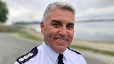 A man smiles towards the camera , he has grey hair and is wearing a white shirt with a black tie, the shirt has black epaulettes on the shoulders with three silver discs. There is a lake behind him.