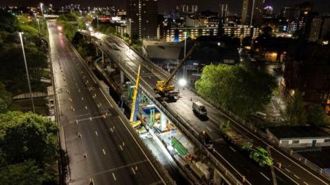 An overhead view of the roadworks on the M8. The shot is taken of night, showing vehicles and cranes on an overpass section, with high-rise buildings on the left and trees on either side of the road, which is devoid of traffic. 