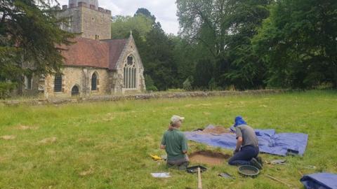 Two people digging in a hole on grass in the foreground of a church