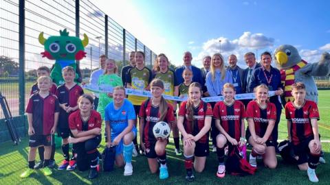 Group of young footballers, pose with mascots and dignitaries on a football pitch