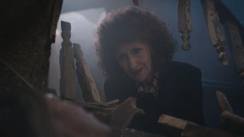 Anita Dobson dressed as Angie Watts in her return to EastEnders. She is wearing a dark, curly wig and is looking down through broken staircase bannisters.