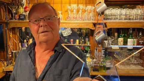 Lawrence Dixon is standing on the left behind a bar. He is wearing a navy shirt and clear framed glasses. He is holding a star shaped award.