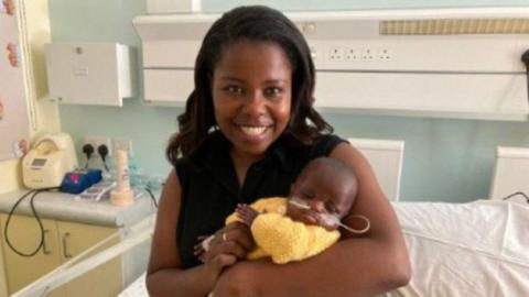 A mother smiles in a hospital ward as she cradles a small baby wearing a yellow knitted cardigan. He has a feeding tube in his nose and his eyes are shut. The mother is wearing a sleeveless black top and has long black hair.