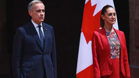 Prime Minister Mark Carney pictured to the left of President Claudia Sheinbaum. He is wearing a navy blue suit with a matching tie, and a white button-up shirt. Sheinbaum is to his right, wearing a red suit jacket and skirt with a traditional Mexican patterned top. The two are standing straight, looking ahead to the right of the camera. Behind Sheinbau, is a Canadian flag on a pole. 