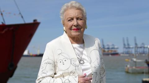 Dame Stephanie Shirley poses infront of the sea in a white suit covered in drawings of children and a Star of David necklace at the unveiling of a memorial to Kindertransport on the quayside at Harwich in 2022