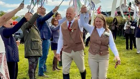 Safron and Nigel walking down a field with people either side holding decorations in the air. The pair wear black boots, beige trousers, brown tweed jacket and white shirts. Nigel holds Safron's hand into air.