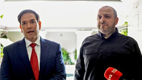 A man in blue suit, white shirt and red tie stands close to a man in a dark jacket.