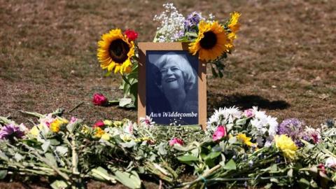 A framed black and white picture of Ann Widdecombe smiling is placed upon a grassy area, surrounded by flowers including sunflowers 