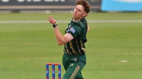 James Hayes, in green and gold kit, bowling alongside blue stumps with red bails