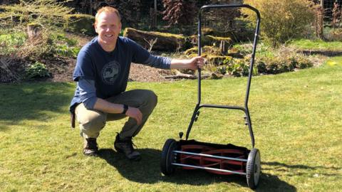 A man crouching next to a push mower on a lawn.