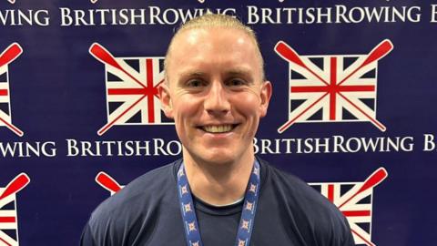 A smiling Ben Stephens wearing his gold medal stands in front of the British Rowing logo.  