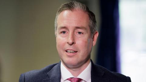 Paul Givan, a man with blue eyes and short grey hair, looks towards the camera.  He is mid-speech and is wearing a navy suit, a white shirt and a pink tie.